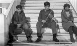 Children, one with violin, in front of 3rd residential school at Onion Lake, Saskatchewan